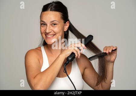 donna sorridente che stira i capelli con piastra su sfondo grigio Foto Stock
