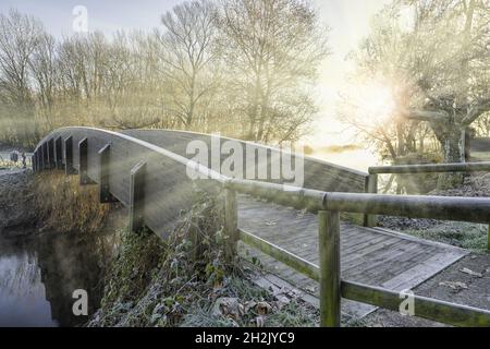 Ponte all'alba in inverno e raggi del sole tra la foschia. Ponte lungo la pista ciclabile pedonale intorno al Lago di Varese, Italia, sul fiume Bardello Foto Stock