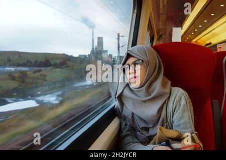 Ritratto di giovane e bella donna musulmana asiatica indossando occhiali e hijab seduto da solo contro la finestra e guardando fuori in un treno in movimento. Foto Stock