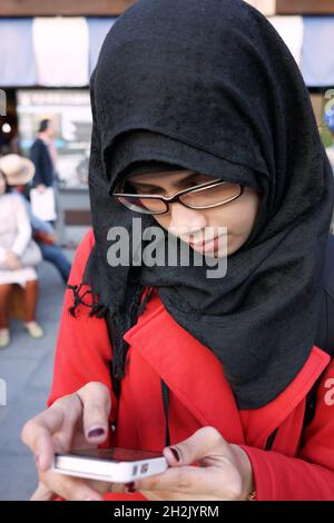 Primo piano ritratto della giovane donna musulmana asiatica che indossa hijab nero e occhiali tenendo e guardando smartphone in strada di Kyoto, Giappone. Foto Stock