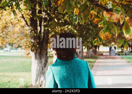 Una ragazza in un cappotto blu con capelli ricci scuri che scorrono su un vicolo autunnale di castagni, vista posteriore Foto Stock