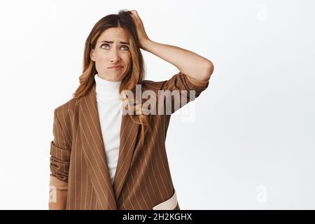 Confuso donna di mezza età che ha problemi, pensiero e testa graffiante ignaro, clueless come risolvere la situazione, in piedi disturbato su bianco Foto Stock