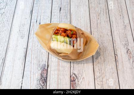 Panino Bao con pollo marinato in salsa dolce e aspre, erba cipollina e pezzi di cetriolo crudo avvolti in carta pergamena marrone Foto Stock