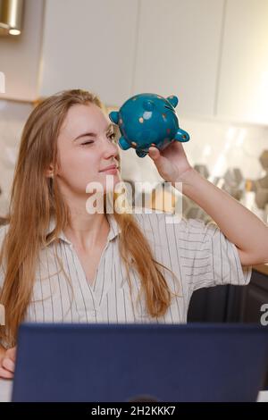 Donna che guarda a vuoto Piggy banca seduta a casa con notebook in cucina, bilancio di famiglia, risparmio personale shopping online, credito Foto Stock