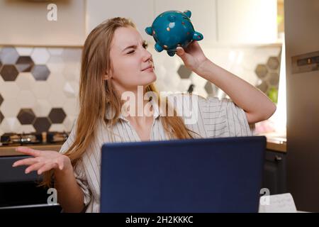 Donna che guarda a vuoto Piggy banca seduta a casa con notebook in cucina, bilancio di famiglia, risparmio personale shopping online, credito Foto Stock