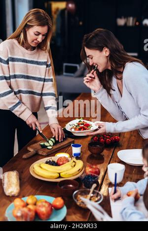 Due giovani donne stanno preparando il pranzo, una sta tagliando le verdure per un'insalata, l'altra sta degustando e la ragazza vicino a lei sta facendo i suoi compiti Foto Stock