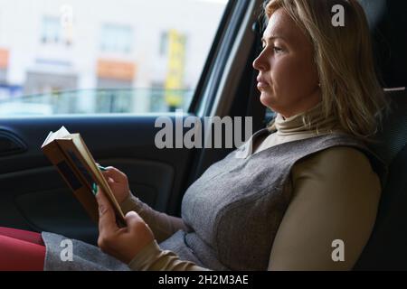 Una donna calma di mezza età legge un libro interessante mentre si siede in una macchina nel traffico. Foto Stock