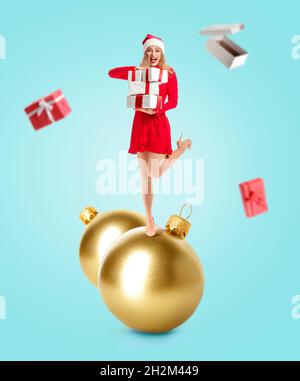 Felice giovane donna in Santa cappello e con regali in piedi su grande palla di Natale su sfondo di colore Foto Stock