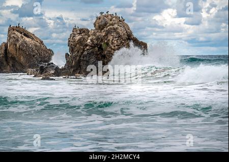 L'acqua dell'oceano spruzzi sulla spiaggia di roccia con il cielo e le nuvole bei. Onda di mare che si tuffa in pietra a riva del mare in inverno. Costa rocciosa con acqua bianca s Foto Stock