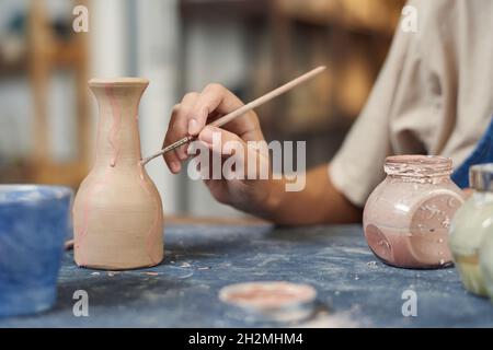 Primo piano di potter non riconoscibile utilizzando il pennello mentre si applica la vernice rosa su vaso di argilla in officina Foto Stock
