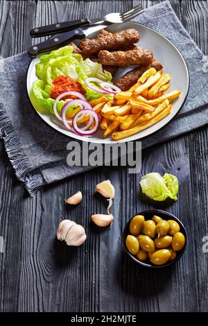 cevapcici, salsicce balcaniche grigliate con patatine fritte, anelli di cipolla rossa, ajvar e foglie di lattuga fresche su un piatto su un tavolo di legno nero con olietto verde Foto Stock