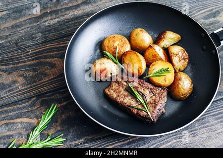 Patate fritte fatte in casa con bistecca di manzo con rosmarino in una padella su un tavolo di legno Foto Stock