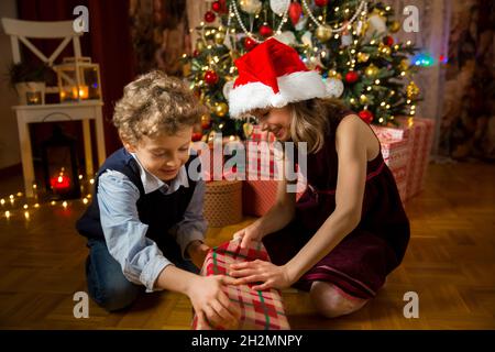 Eccitati i bambini carini felicemente sorridenti, regali di Natale di apertura. Soggiorno splendidamente decorato con luci e albero di Natale. I bambini si divertono Foto Stock