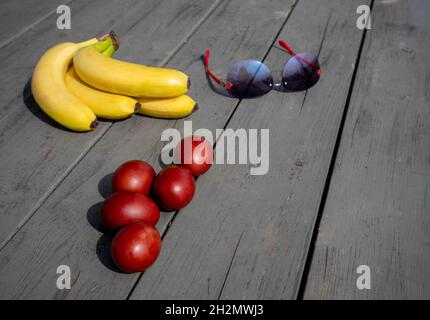 Mescola la composizione con uova di pasqua dipinte, mazzo di banane mature e occhiali da sole giacenti su una vecchia superficie di legno Foto Stock