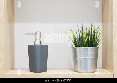 Dispenser di sapone e casalinga artificiale in pentola decorativa in metallo su ripiano in legno in bagno. Dettagli interni Foto Stock