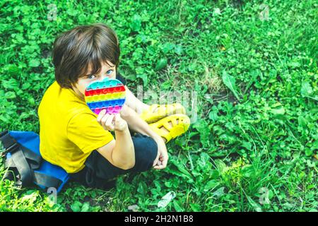 Carino ragazzo del preschooler che gioca con il giocattolo flessibile sensoriale del fidget del Pop IT seduto sull'erba all'aperto. Attività per bambini, sviluppo mentale. Selettivo Foto Stock