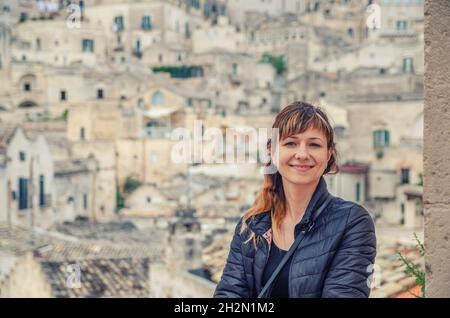 Giovane bella donna viaggiatore con giacca nera guardando la macchina fotografica, sorriso e posa in Sassi di Matera centro storico Sasso Caveoso con vecchio Foto Stock