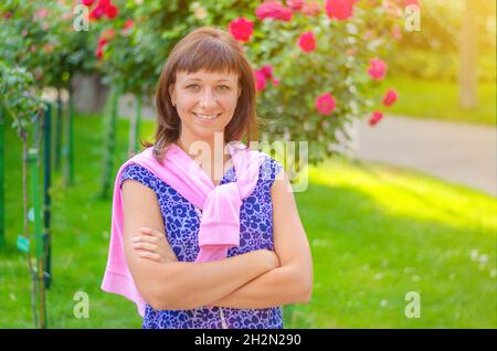 Giovane bella ragazza caucasica con capelli scuri e camicia fiorita guardando la macchina fotografica, posa, incrociando le braccia e sorriso in giardino con fiori e verde t Foto Stock