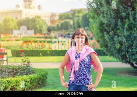 Giovane bella ragazza caucasica con camicia fiorita guardando la macchina fotografica, posa e sorriso nel parco giardino Volksgarten con fiori nella città storica di Vienna Foto Stock