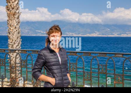 Giovane bella donna viaggiatore guardando la macchina fotografica, sorridere, posare e tenere le mani in tasca sul lungomare Falcomata a Reggio Calabria città, S. Foto Stock