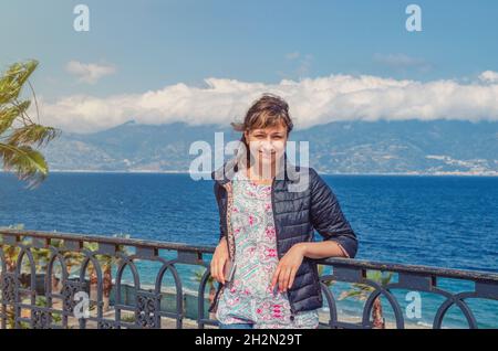 Giovane bella donna viaggiatore guardando macchina fotografica, sorriso e posa sul lungomare Falcomata a Reggio Calabria città, stretto di Messina e Sicil Foto Stock