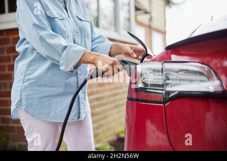 Primo piano della donna che collega il cavo di ricarica a un'auto elettrica a emissioni zero ecologica a casa Foto Stock