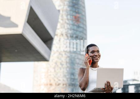 Sorridente freelance donna che tiene il laptop mentre parla con lo smartphone Foto Stock