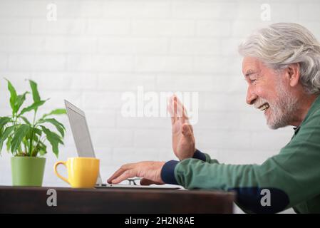 Uomo anziano sorridente che agitava la mano durante le videochiamate attraverso il laptop a casa Foto Stock