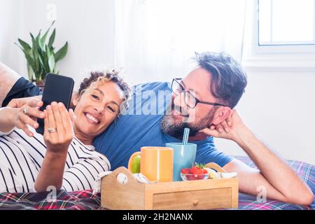 Coppia sorridente che condivide il telefono cellulare sul letto di casa Foto Stock