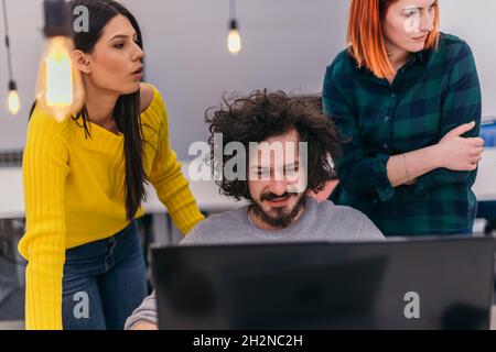 Due giovani donne d'affari accanto a un collega maschile mentre spiega le nuove attività online su un computer in un ambiente di ufficio confortevole. Foto Stock