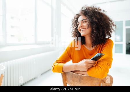 Sorridente donna afroica che tiene lo smartphone mentre si siede sulla sedia a casa Foto Stock