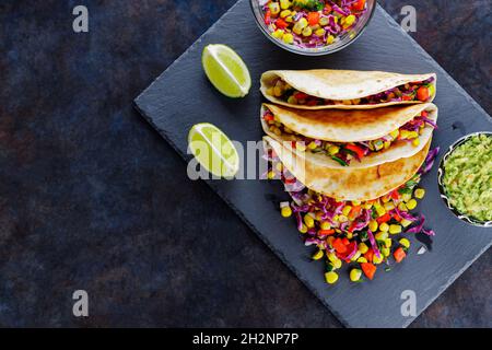 Tacos vegetariani con verdure varie, guacamole e lime a fette su sfondo scuro. Tacos con mais dolce, cavolo viola e pepe su un baro di ardesia Foto Stock