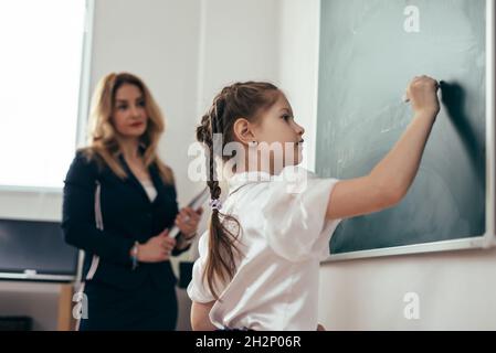 Schoolgirl first-grader che scrive sulla lavagna. Lezione di scuola insegnante e allievo Foto Stock