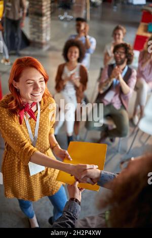 La giovane donna in carica che si riunisce in un'atmosfera piacevole sta scuotendo le mani con un collega dopo aver ricevuto le congratulazioni per il merito al lavoro. Impiegare Foto Stock