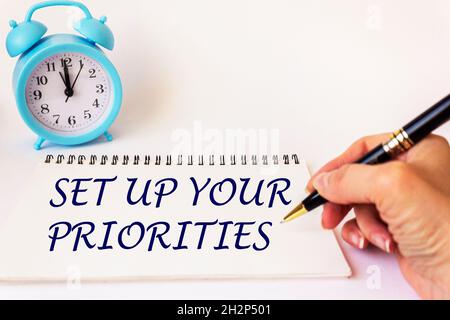 La mano di una donna scrive il testo su un blocco note impostare le vostre priorità. Sfondo bianco con orologio. Affari Foto Stock