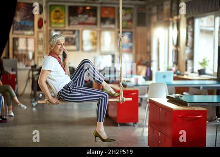 Una donna anziana che si rilassa in un'altalena in un'atmosfera piacevole in ufficio e parla con uno smartphone. Dipendenti, ufficio, lavoro Foto Stock