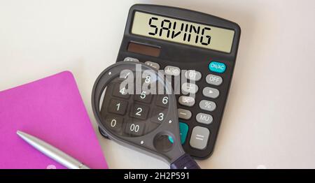 Calcolatrice con risparmio sul display su sfondo bianco, vicino a una lente di ingrandimento, blocco note e penna Foto Stock