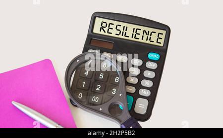 Resilienza, testo scritto su una calcolatrice, accanto ad un blocco note, una lente d'ingrandimento e una penna su sfondo bianco. Foto Stock