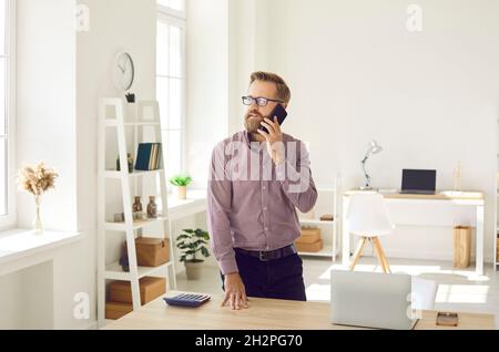Uomo d'affari serio in piedi da ufficio scrivania con laptop e fare chiamata sul telefono cellulare Foto Stock