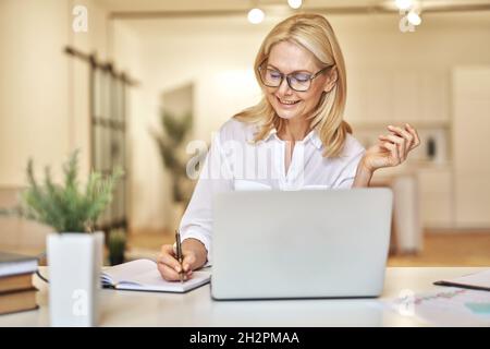 Attraente donna d'affari matura sorridente e prendere appunti mentre si fa una videochiamata online utilizzando un computer portatile, seduto alla sua scrivania in ufficio Foto Stock