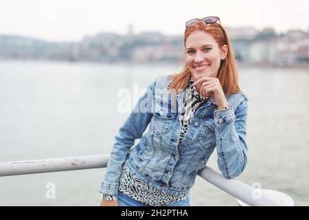 Giovane allegra rossa caucasica donna naturale guardando in macchina fotografica a ringhiera sul fiume Foto Stock