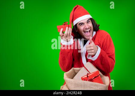 L'uomo entusiasta vestito come Babbo Natale mostra un piccolo regalo rosso preso da un sacco pieno di regali ed esprime la sua aprecciazione con il pancino in su Foto Stock