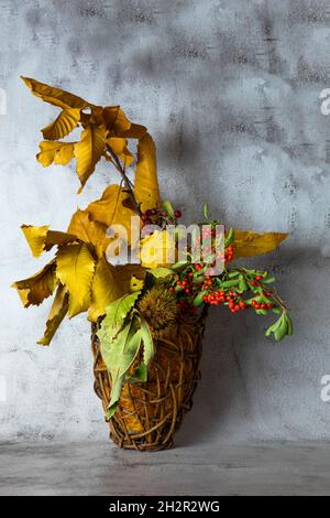 Autunno ikebana.Fall foglie di castagno giallo, rami con rosso selvatico e frutti di bosco di arancia in un cestino di vimini Foto Stock