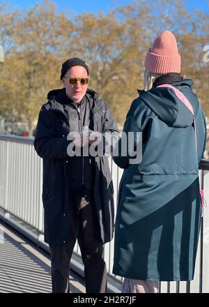 Un uomo sta rompendo il sandwich per condividere con una donna in piedi faccia a faccia con lui. Si ergono su un ponte e ci sono alberi con foglie d'autunno gialle. Foto Stock