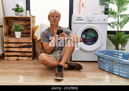 Giovane bionda che fa lavanderia usando smartphone pensando concentrato sul dubbio con il dito sul mento e guardando in su domandandosi Foto Stock