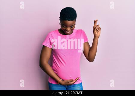 Giovane afroamericana che aspetta un bambino, toccando ventre incinta gesturante dito attraversato sorridendo con speranza e occhi chiusi. Fortuna e superstiti Foto Stock