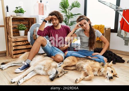 Giovane coppia ispanica che fa la lavanderia con i cani preoccupati e sollecitati circa un problema con la mano sulla fronte, nervoso e ansioso per la crisi Foto Stock