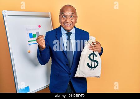 Uomo latino di Medio Evo che indossa il vestito di affari che tiene i dollari sacchetto urlando orgoglioso, celebrando la vittoria ed il successo molto eccitato con le armi sollevate Foto Stock