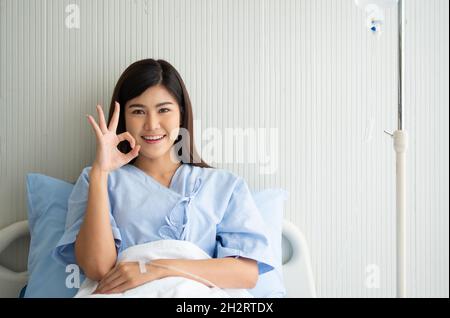 Felice paziente asiatico femmina sorridente, si trova sul letto, e mostra un simbolo ok per mostrare fiducia nel trattamento. Concetto di credere nel trattamento e insur Foto Stock