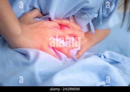 Paziente asiatico infelice donna seduta sul letto d'ospedale e tenuta sullo stomaco sofferenza. Dolore addominale che deriva da mestruazioni, diarrea, o in Foto Stock